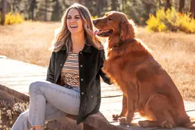 Woman sitting with dog on wooden walkway in field.